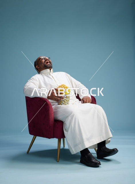 بورتريه لرجل  عربي سعودي خليجي يرتدي الثوب الابيض  يجلس على كرسي مريح, الاستمتاع بتناول  الفشار , خلفيه زرقاء
