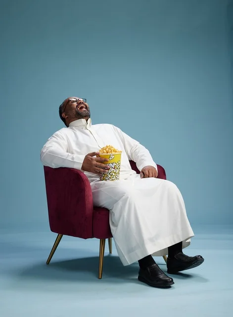 A portrait of an Arab Saudi Gulf man wearing a white thobe sitting on a comfortable chair, enjoying eating popcorn, with a blue background.