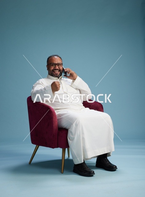 بورتريه لرجل سعودي بالزي التقليدي جالس ويتحدث في الهاتف, اجراء مكامله هاتفيه عبر الجوال , خلفيه زرقاء