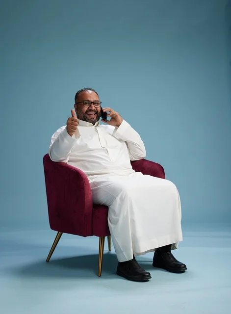 A portrait of a Saudi man in traditional attire sitting and talking on the phone, conducting a phone conversation via mobile, with a blue background.