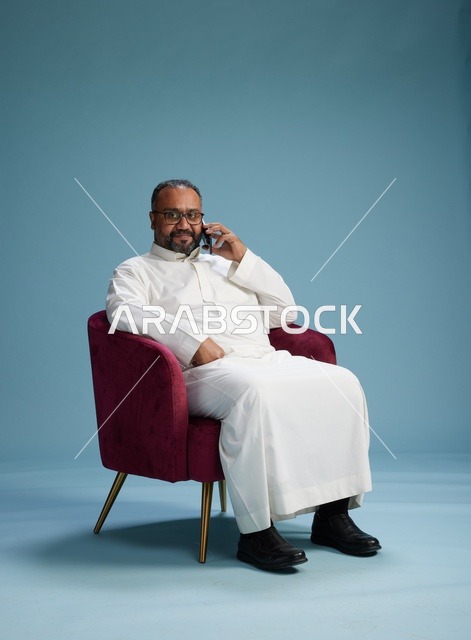 بورتريه لرجل سعودي بالزي التقليدي جالس ويتحدث في الهاتف, اجراء مكامله هاتفيه عبر الجوال , خلفيه زرقاء