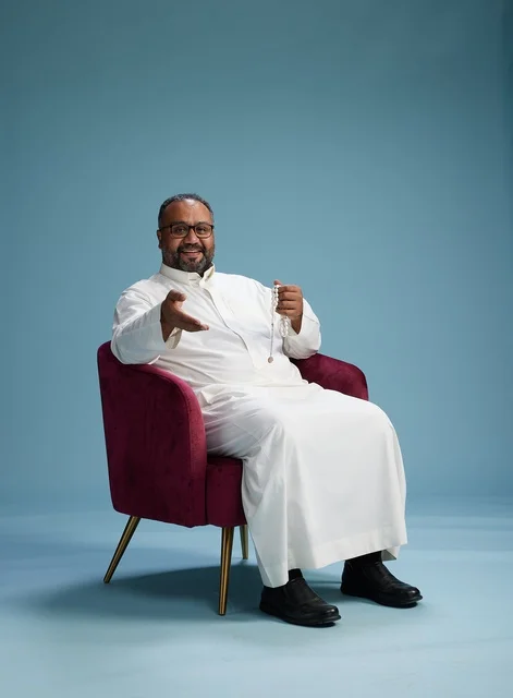 بورتريه لرجل  عربي سعودي  خليجي يرتدي الثوب الابيض يجلس على كرسي احمر,  إيماءة تعبيرية متنوعه, خلفيه زرقاء