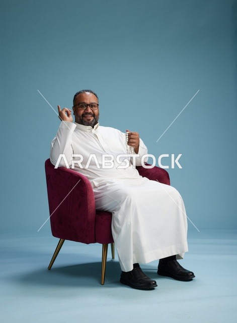 بورتريه لرجل  عربي سعودي  خليجي يرتدي الثوب الابيض يجلس على كرسي احمر,  إيماءة تعبيرية متنوعه, خلفيه زرقاء
