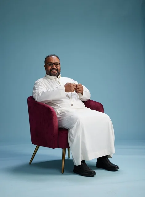 A portrait of a Saudi Arabian Gulf man wearing a white thobe sitting on a red chair, with various expressive gestures, against a blue background.