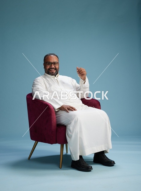 بورتريه لرجل  عربي سعودي  خليجي يرتدي الثوب الابيض يجلس على كرسي احمر,  إيماءة تعبيرية متنوعه, خلفيه زرقاء
