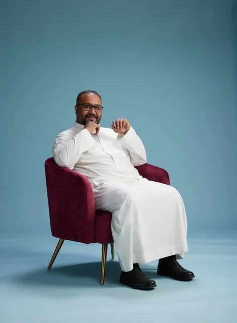A portrait of a Saudi Arabian Gulf man wearing a white thobe sitting on a red chair, with various expressive gestures, against a blue background.