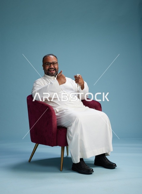 بورتريه لرجل  عربي سعودي  خليجي يرتدي الثوب الابيض يجلس على كرسي احمر,  إيماءة تعبيرية متنوعه, خلفيه زرقاء