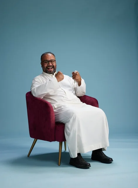 A portrait of a Saudi Arabian Gulf man wearing a white thobe sitting on a red chair, with various expressive gestures, against a blue background.
