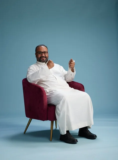 A portrait of a Saudi Arabian Gulf man wearing a white thobe sitting on a red chair, with various expressive gestures, against a blue background.