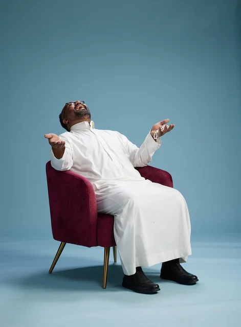 A portrait of a Saudi Arabian Gulf man wearing a white thobe sitting on a red chair, with various expressive gestures, against a blue background.