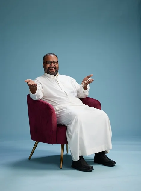 A portrait of a Saudi Arabian Gulf man wearing a white thobe sitting on a red chair, with various expressive gestures, against a blue background.