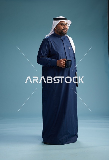 رجل سعودي مبتسم يرتدي ثوب كحلي وغتره بيضاء يحمل كوب من القهوة ولابتوب,  الذهاب الى العمل, كامل الجسم, خلفيه زرقاء