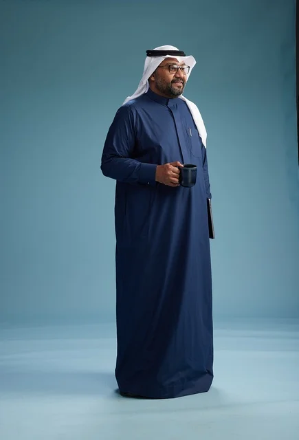 A smiling Saudi man wearing a navy blue thobe and a white ghutrah is holding a cup of coffee and a laptop, going to work, full body, blue background.