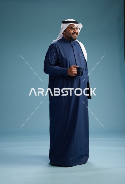 رجل سعودي مبتسم يرتدي ثوب كحلي وغتره بيضاء يحمل كوب من القهوة ولابتوب,  الذهاب الى العمل, كامل الجسم, خلفيه زرقاء