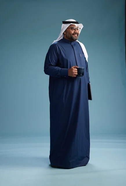 A smiling Saudi man wearing a navy blue thobe and a white ghutrah is holding a cup of coffee and a laptop, going to work, full body, blue background.