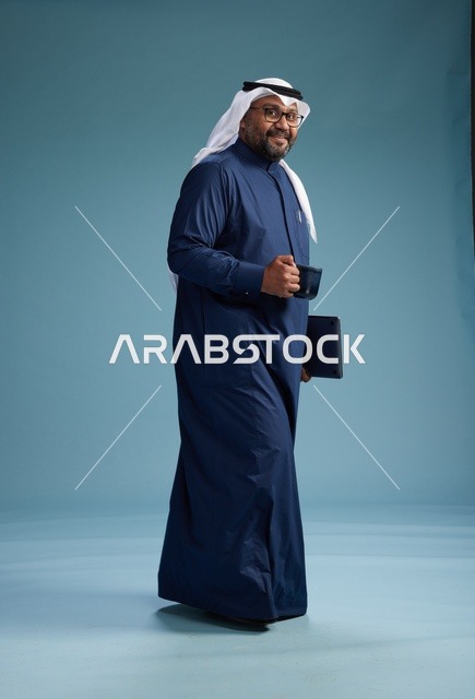 رجل سعودي مبتسم يرتدي ثوب كحلي وغتره بيضاء يحمل كوب من القهوة ولابتوب,  الذهاب الى العمل, كامل الجسم, خلفيه زرقاء