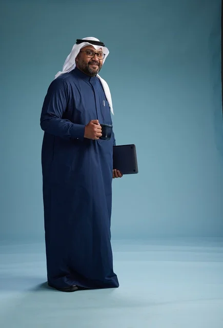 A smiling Saudi man wearing a navy blue thobe and a white ghutrah is holding a cup of coffee and a laptop, going to work, full body, blue background.