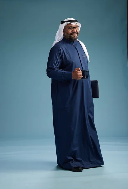 A smiling Saudi man wearing a navy blue thobe and a white ghutrah is holding a cup of coffee and a laptop, going to work, full body, blue background.