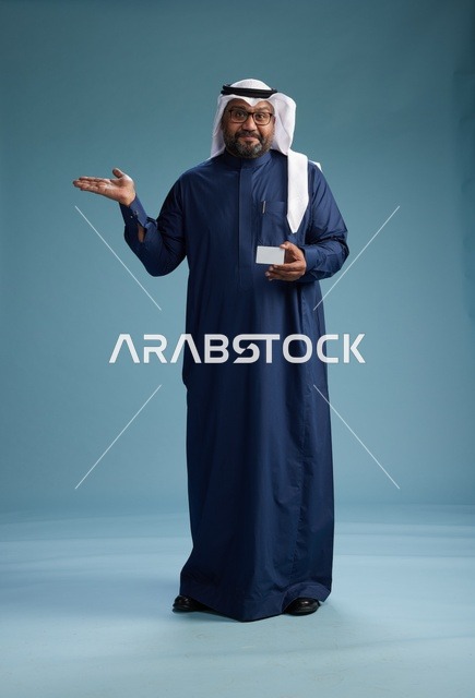 بورتريه لرجل سعودي يرتدي ثوب كحلي وغتره بيضاء يحمل بطاقة ائتمان ويقف, الجسم بالكامل, بطاقه بيضاء فارغه, خلفيه زرقاء