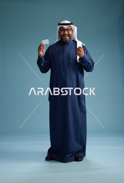 بورتريه لرجل سعودي يرتدي ثوب كحلي وغتره بيضاء يحمل بطاقة ائتمان ويقف, الجسم بالكامل, بطاقه بيضاء فارغه, خلفيه زرقاء