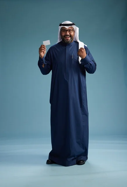 A portrait of a Saudi man wearing a navy thobe and a white ghutrah, holding a credit card and standing, full body, with a blank white card, against a blue background.