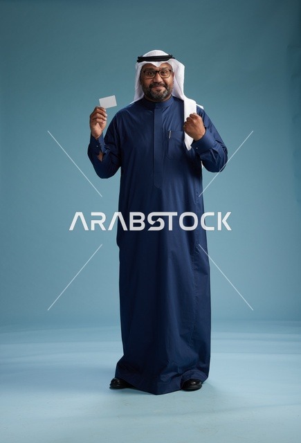 بورتريه لرجل سعودي يرتدي ثوب كحلي وغتره بيضاء يحمل بطاقة ائتمان ويقف, الجسم بالكامل, بطاقه بيضاء فارغه, خلفيه زرقاء