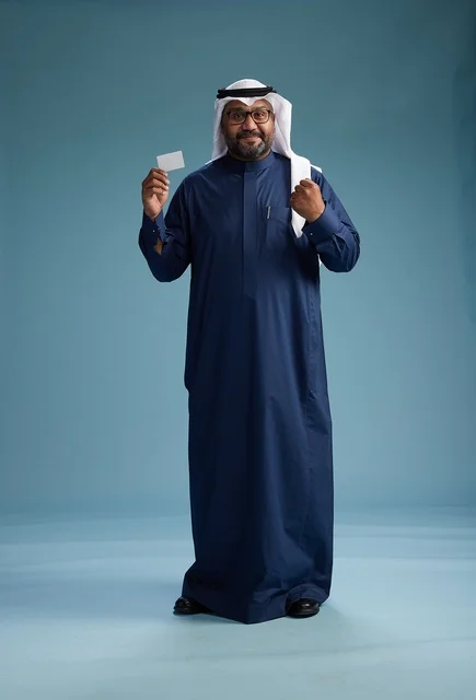 A portrait of a Saudi man wearing a navy thobe and a white ghutrah, holding a credit card and standing, full body, with a blank white card, against a blue background.