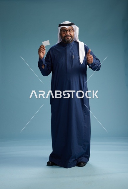 بورتريه لرجل سعودي يرتدي ثوب كحلي وغتره بيضاء يحمل بطاقة ائتمان ويقف, الجسم بالكامل, بطاقه بيضاء فارغه, خلفيه زرقاء