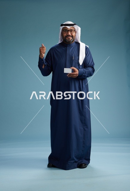 بورتريه لرجل سعودي يرتدي ثوب كحلي وغتره بيضاء يحمل بطاقة ائتمان ويقف, الجسم بالكامل, بطاقه بيضاء فارغه, خلفيه زرقاء