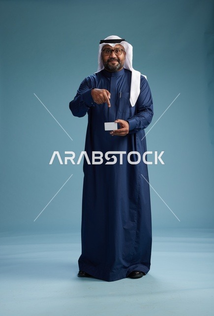 بورتريه لرجل سعودي يرتدي ثوب كحلي وغتره بيضاء يحمل بطاقة ائتمان ويقف, الجسم بالكامل, بطاقه بيضاء فارغه, خلفيه زرقاء