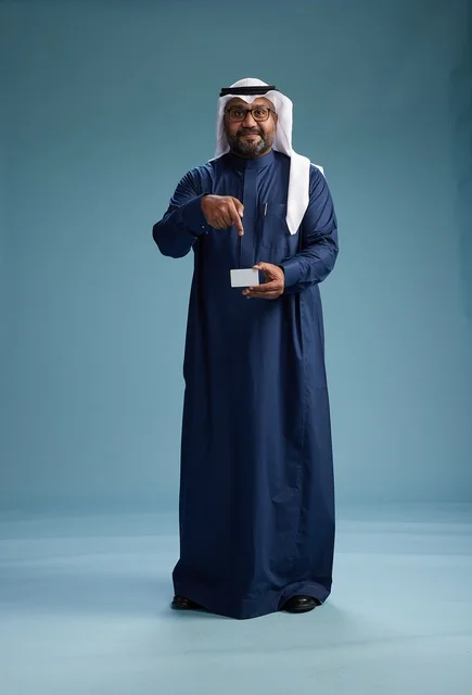 بورتريه لرجل سعودي يرتدي ثوب كحلي وغتره بيضاء يحمل بطاقة ائتمان ويقف, الجسم بالكامل, بطاقه بيضاء فارغه, خلفيه زرقاء