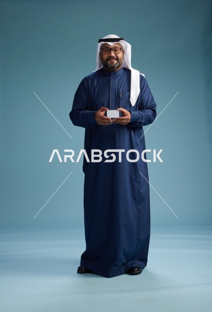 بورتريه لرجل سعودي يرتدي ثوب كحلي وغتره بيضاء يحمل بطاقة ائتمان ويقف, الجسم بالكامل, بطاقه بيضاء فارغه, خلفيه زرقاء