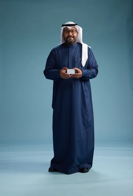 A portrait of a Saudi man wearing a navy thobe and a white ghutrah, holding a credit card and standing, full body, with a blank white card, against a blue background.
