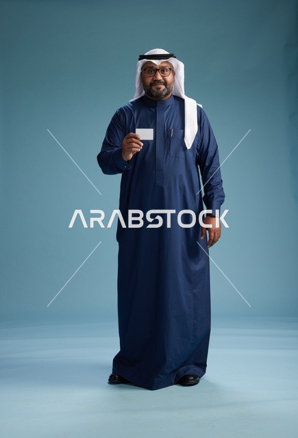 بورتريه لرجل سعودي يرتدي ثوب كحلي وغتره بيضاء يحمل بطاقة ائتمان ويقف, الجسم بالكامل, بطاقه بيضاء فارغه, خلفيه زرقاء