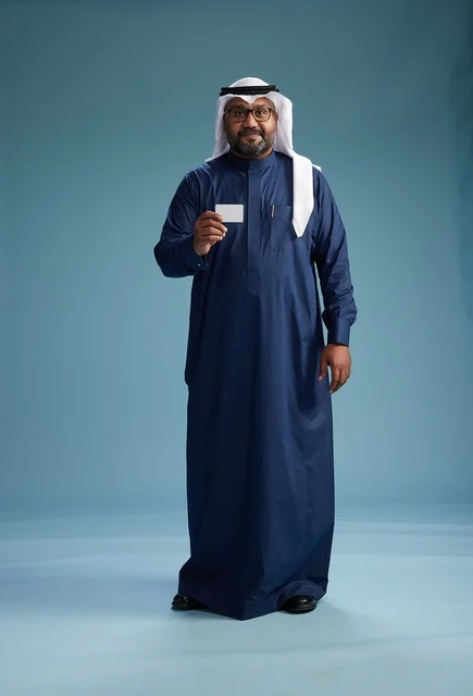 A portrait of a Saudi man wearing a navy thobe and a white ghutrah, holding a credit card and standing, full body, with a blank white card, against a blue background.
