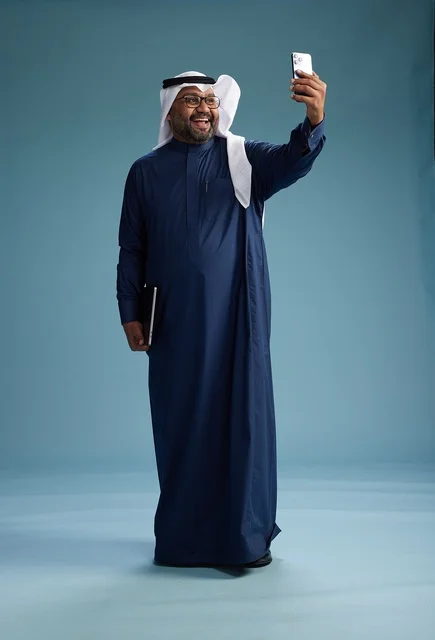A Saudi man wearing a blue thobe and a white ghutrah is smiling and taking a selfie using a smartphone, making a video call via mobile, full body, with a blue background.