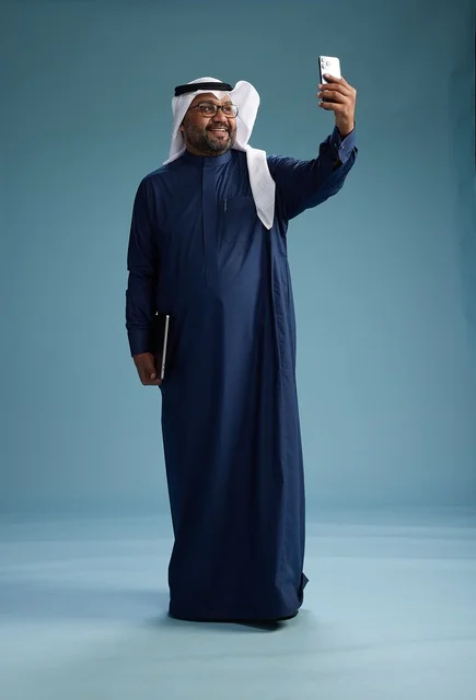 A Saudi man wearing a blue thobe and a white ghutrah is smiling and taking a selfie using a smartphone, making a video call via mobile, full body, with a blue background.