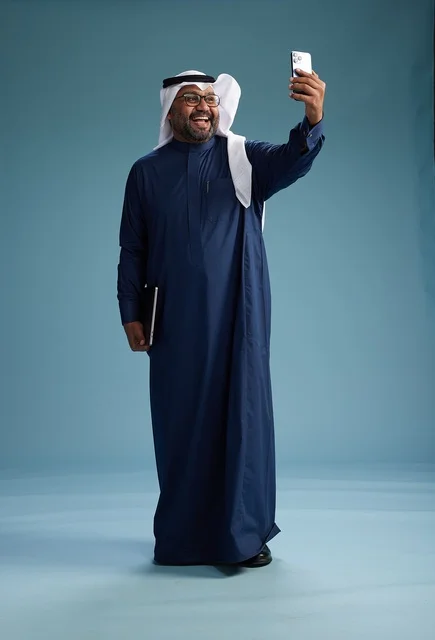 A Saudi man wearing a blue thobe and a white ghutrah is smiling and taking a selfie using a smartphone, making a video call via mobile, full body, with a blue background.
