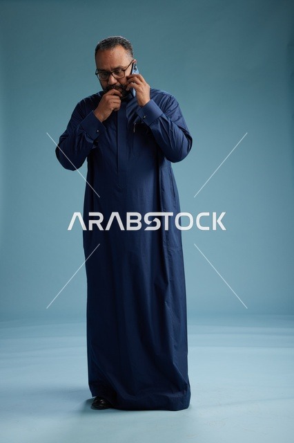 بورتريه لرجل سعودي يرتدي ثوب كحلي في مكالمة هاتفية, دردشه ومحادثات عبر الجوال, الوقوف بالطول الكامل, خلفيه زرقاء
