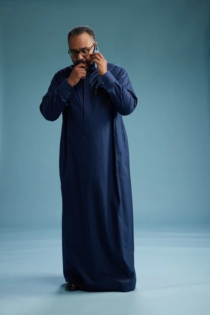 A portrait of a Saudi man wearing a navy blue thobe on a phone call, chatting and conversing via mobile, standing full-length, with a blue background.