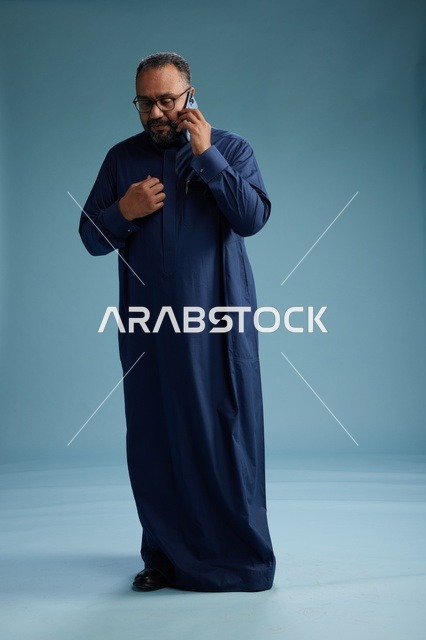 بورتريه لرجل سعودي يرتدي ثوب كحلي في مكالمة هاتفية, دردشه ومحادثات عبر الجوال, الوقوف بالطول الكامل, خلفيه زرقاء