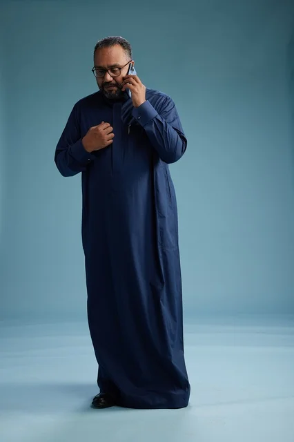 A portrait of a Saudi man wearing a navy blue thobe on a phone call, chatting and conversing via mobile, standing full-length, with a blue background.