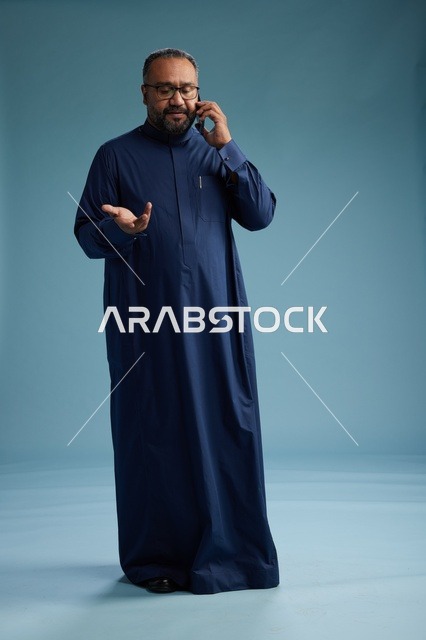 بورتريه لرجل سعودي يرتدي ثوب كحلي في مكالمة هاتفية, دردشه ومحادثات عبر الجوال, الوقوف بالطول الكامل, خلفيه زرقاء