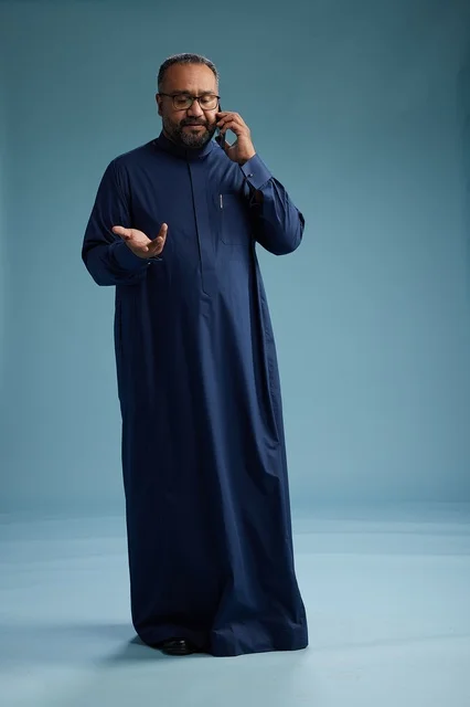 A portrait of a Saudi man wearing a navy blue thobe on a phone call, chatting and conversing via mobile, standing full-length, with a blue background.