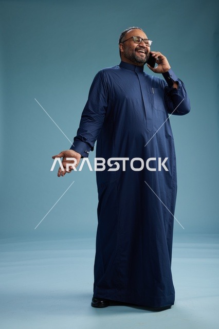 بورتريه لرجل سعودي يرتدي ثوب كحلي في مكالمة هاتفية, دردشه ومحادثات عبر الجوال, الوقوف بالطول الكامل, خلفيه زرقاء