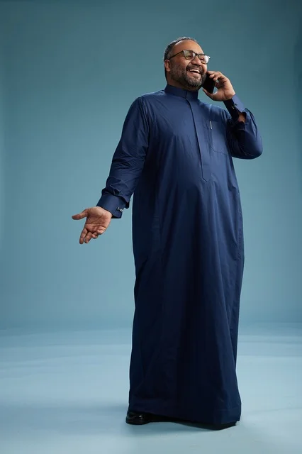 A portrait of a Saudi man wearing a navy blue thobe on a phone call, chatting and conversing via mobile, standing full-length, with a blue background.