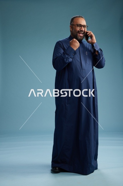 A portrait of a Saudi man wearing a navy blue thobe on a phone call, chatting and conversing via mobile, standing full-length, with a blue background.