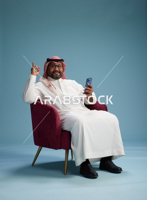 A portrait of a Saudi man wearing a white thobe and a shemagh sitting on a comfortable chair using a mobile phone and gesturing while talking, with a blue background.