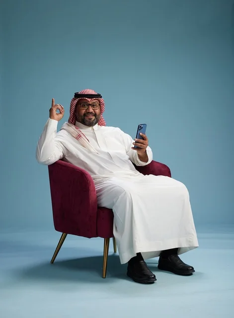 A portrait of a Saudi man wearing a white thobe and a shemagh sitting on a comfortable chair using a mobile phone and gesturing while talking, with a blue background.