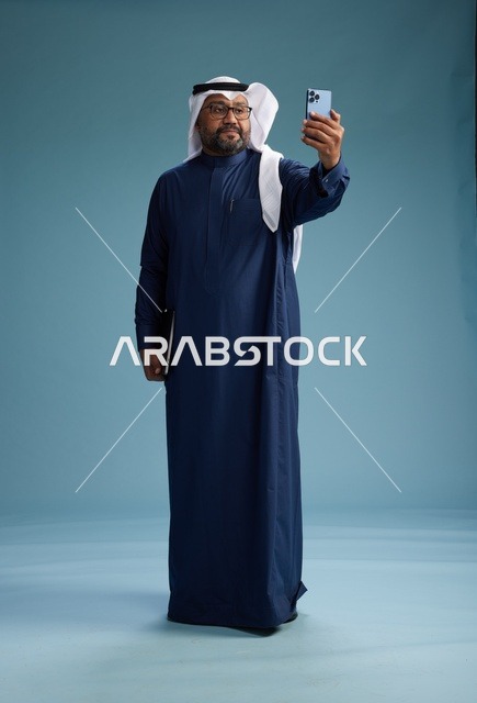 رجل سعودي برتدي ثوب ازرق وغتره بيضاء يبتسم ويلتقط صورة سيلفي باستخدام الهاتف الذكي, اجراء مكاملة فيديو عبر الجوال, كامل الجسم, خلفيه زرقاء